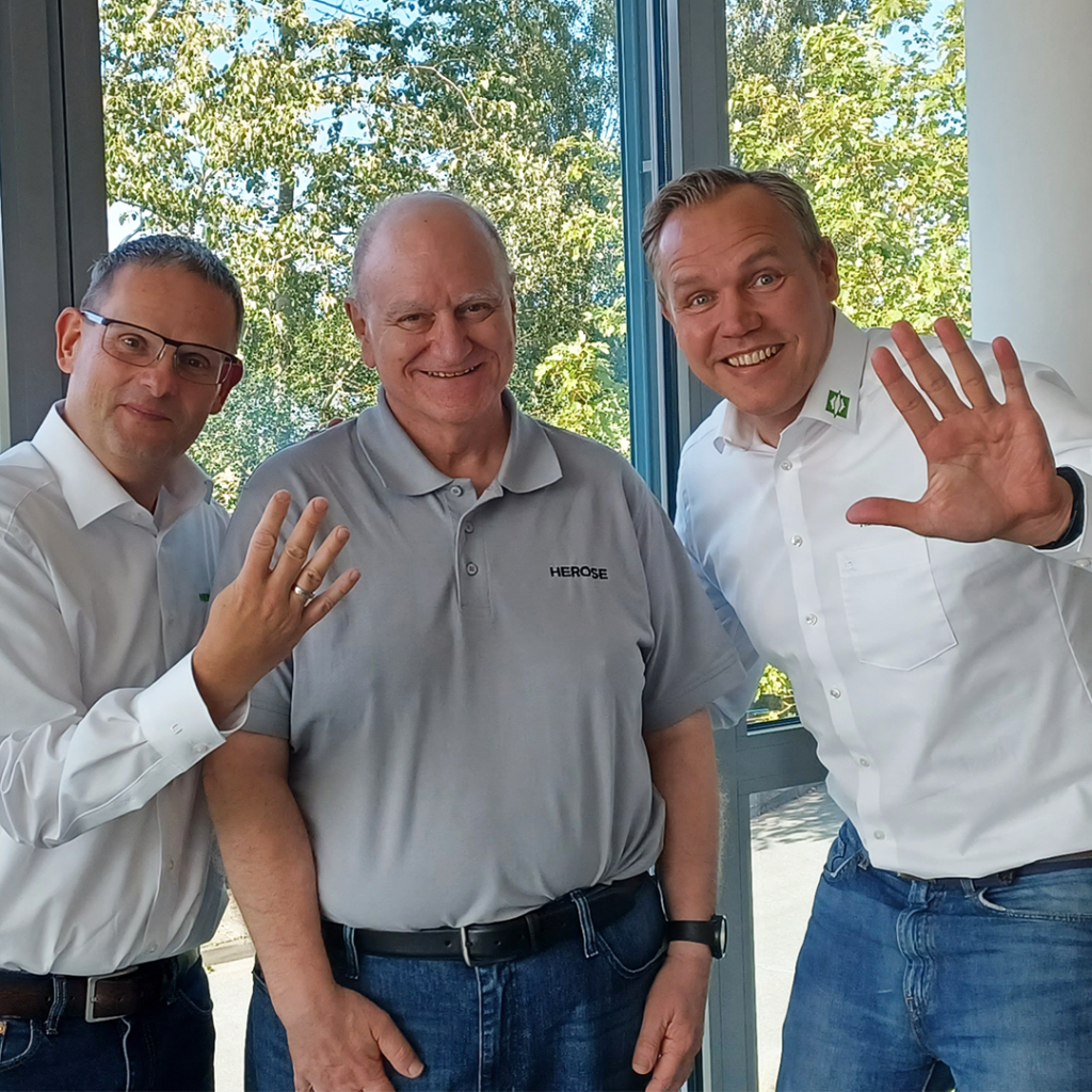 Doug from Mack stands with Dirk and Jens from HEROSE. Dirk holds up four fingers and Jens holds up five. They are smiling.