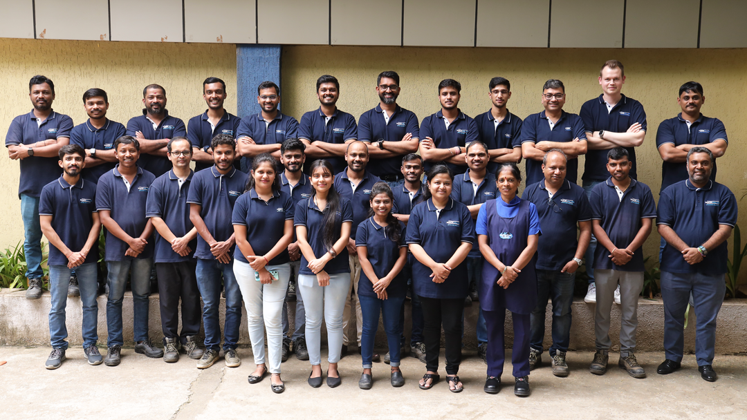 The Mack Valves India team stand in three rows. They are all wearing their Mack Valves uniform and smiling at the camera. They are standing outside and in front of a beige wall of the Mack Valves building. 